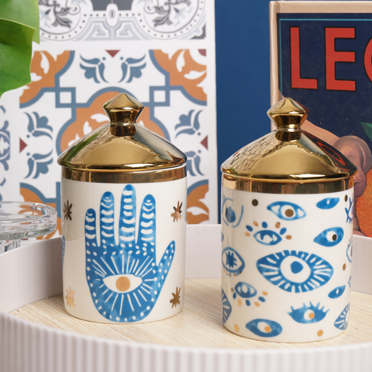 Two ceramic jars with geometric and floral patterns, featuring a gold lid and handle, displayed on a table with a decorative object in the background.